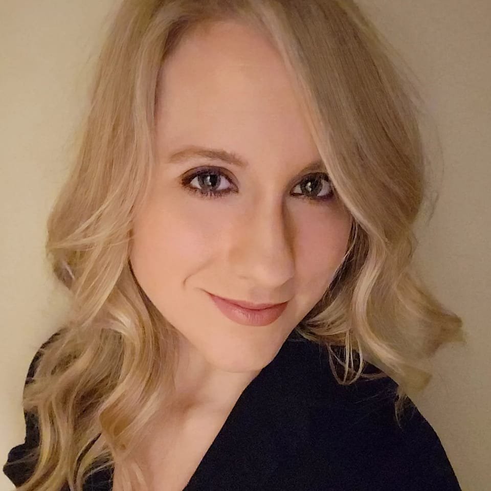 Miss Lenore headshot. Wearing black top in front of beige wall