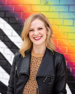Tap, jazz, & musical theatre dance teacher posing outside in a brown shirt and leather jacket with a rainbow and black and white striped background