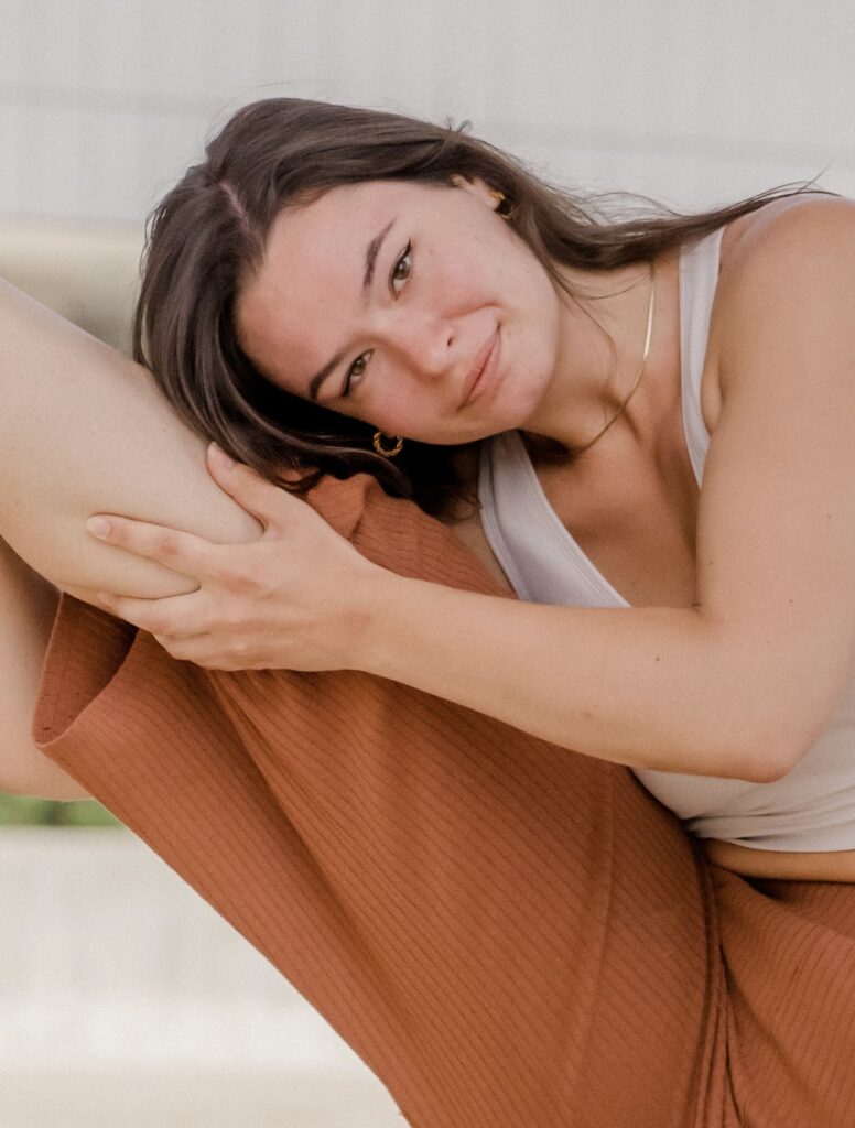 Miss Becca headshot. Orange pants and white tank top holding leg up